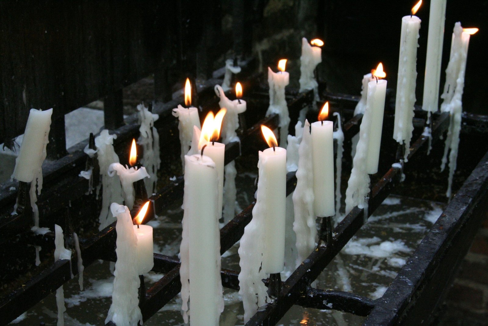 An arrangement of multiple rows of both burning and melted white pillar candles atop a black, metal altar.